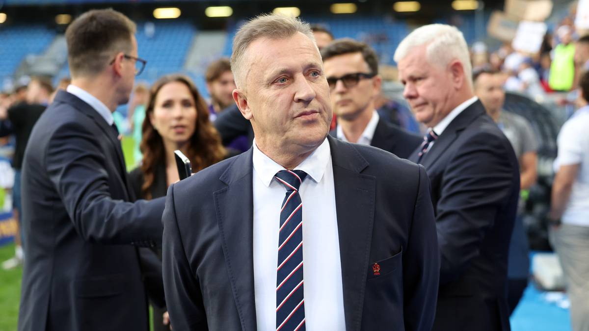 Mężczyzna w garniturze i krawacie, stojący na tle stadionu.