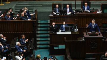 Posłowie zasiadają w ławach sejmowych, niekt&oacute;rzy stoją przy m&oacute;wnicy, trwają obrady.