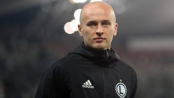 Łysy mężczyzna w czarnej kurtce sportowej z herbem Legii Warszawa.