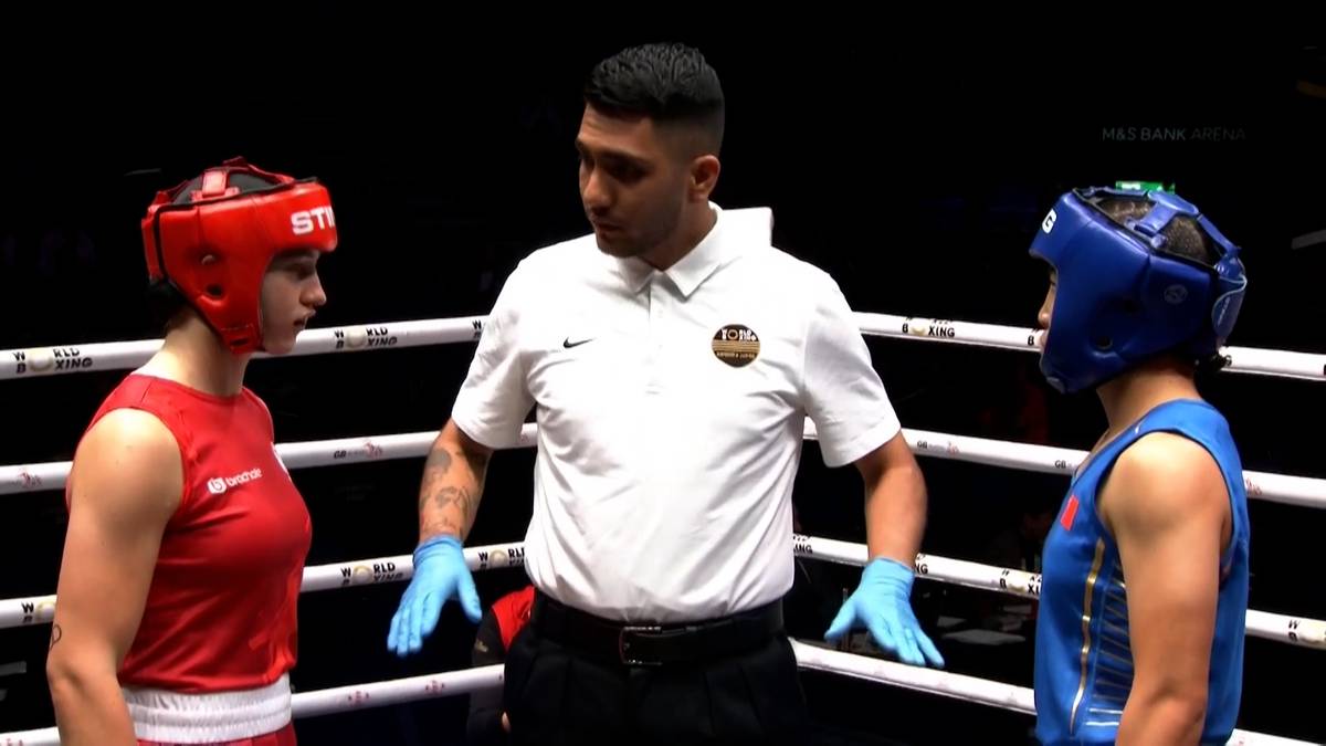 Dwie kobiety w kaskach bokserskich i rękawicach stoją naprzeciwko siebie na ringu bokserskim, z sędzią pomiędzy nimi.