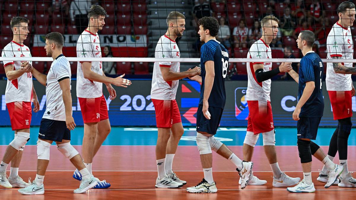 Dw&oacute;ch zawodnik&oacute;w siatk&oacute;wki podaje sobie ręce na środku boiska, podczas meczu. W tle widać innych graczy i kibic&oacute;w na trybunach.