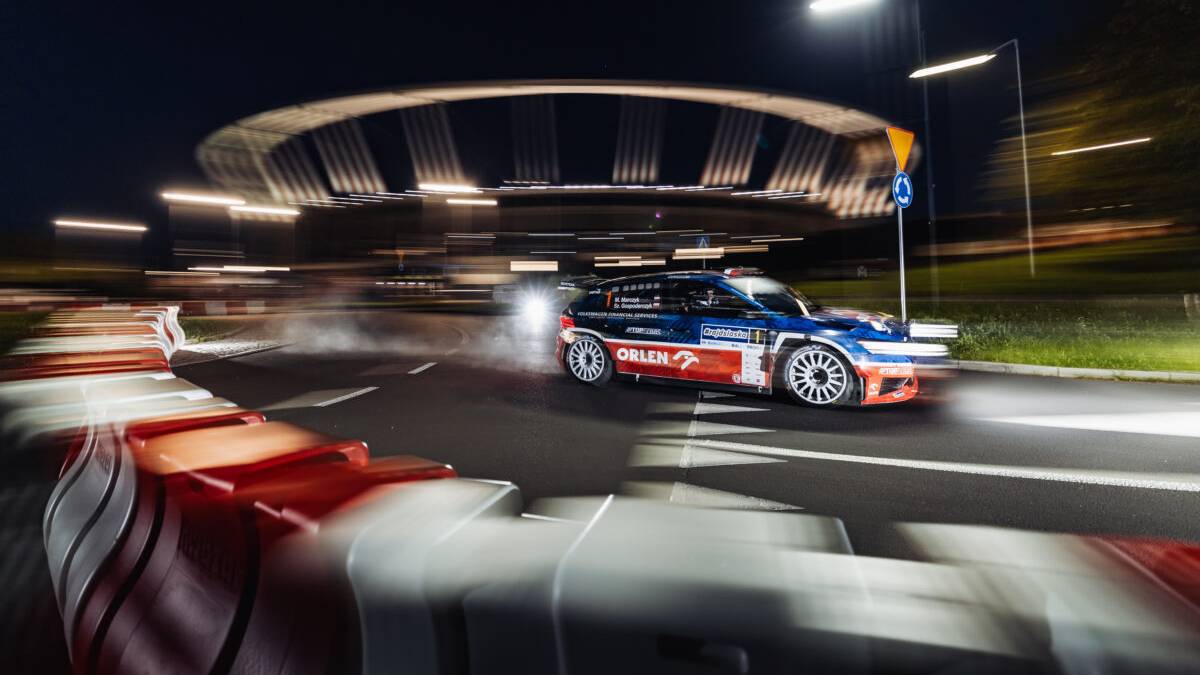 Samochód rajdowy z logo "ORLEN" w ruchu na nocnym odcinku specjalnym, z rozmyciem wskazującym na dużą prędkość. W tle widoczna modernistyczna konstrukcja i światła.
