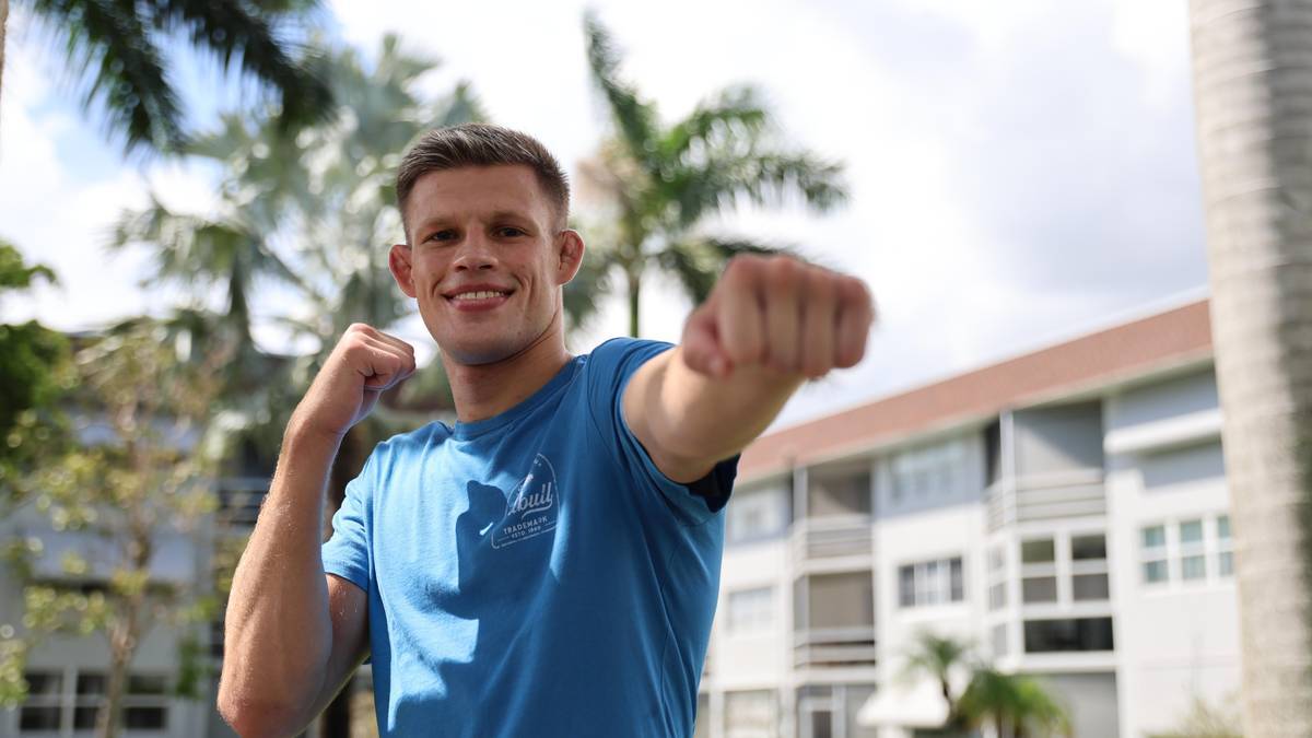 Szczupły mężczyzna w niebieskiej koszulce robi "perkusję" pięściami na tle budynków i palm.