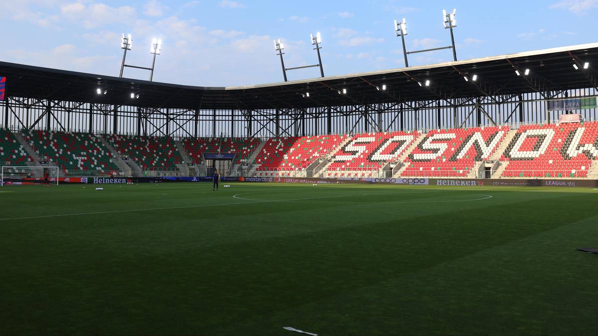 Widok na stadion piłkarski z zieloną murawą i rzędami czerwono-zielonych krzesełek. Na trybunach widnieje napis "SOSNOWIEC".