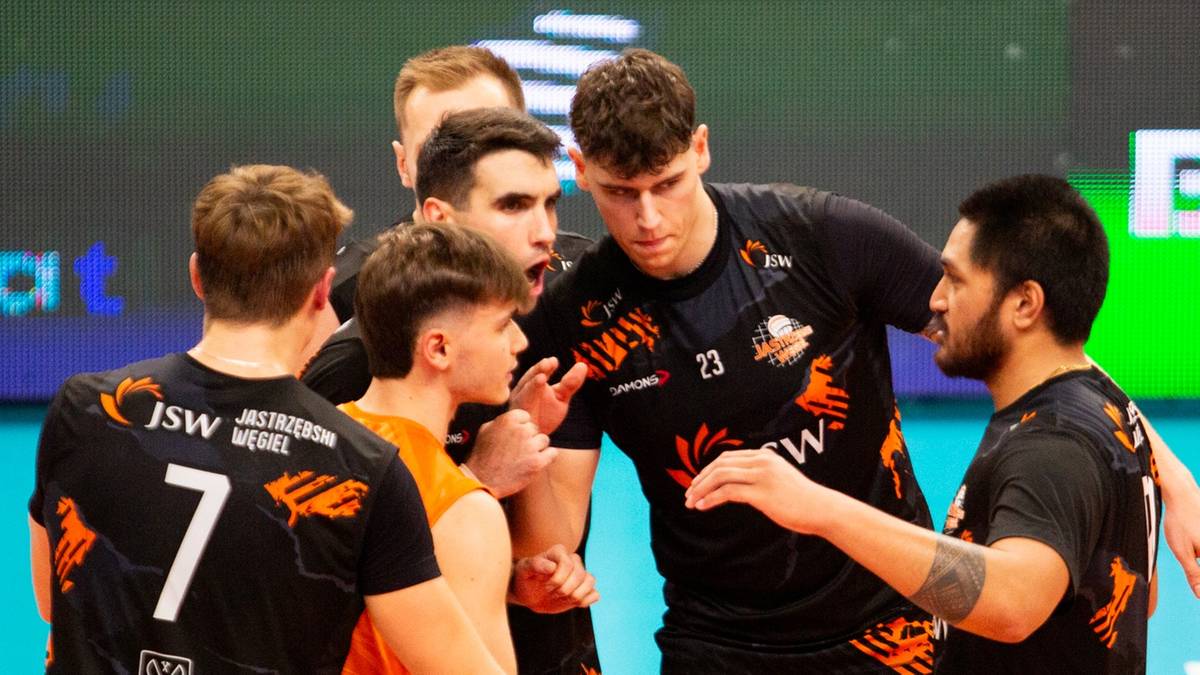 Grupa siatkarzy Jastrzębskiego Węgla w strojach sportowych otacza jednego z nich, słuchając jego słów podczas przerwy w meczu.