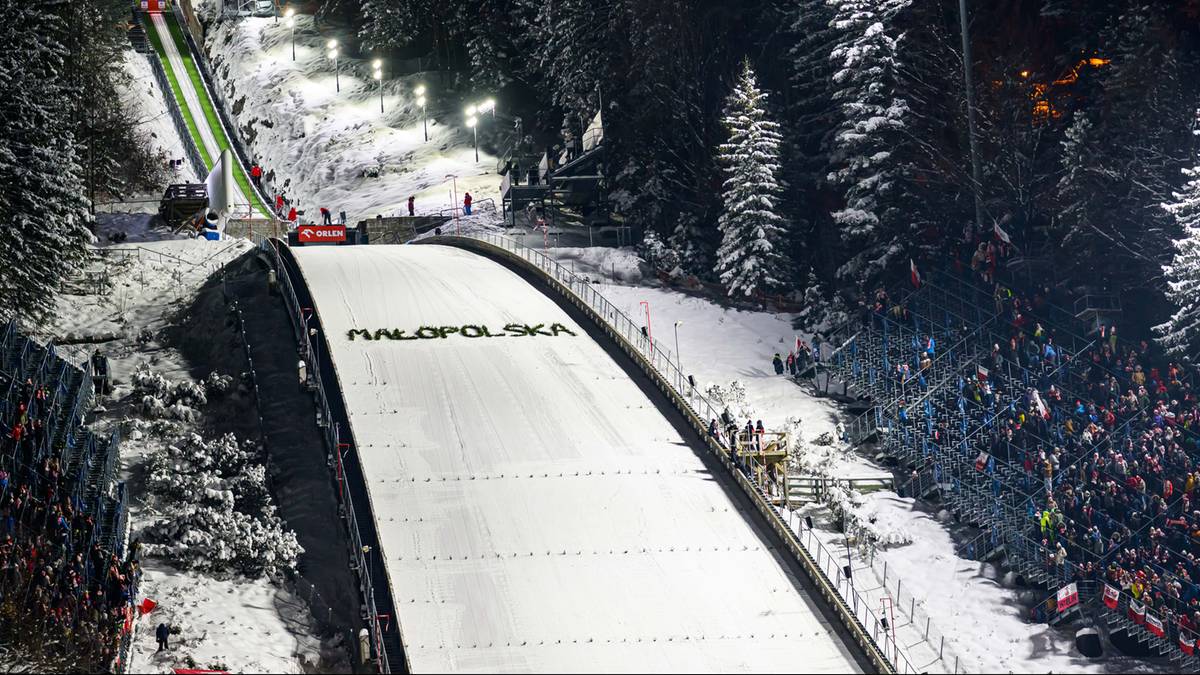 Oświetlona skocznia narciarska nocą, z widownią wypełnioną ludźmi po prawej stronie i lasem iglastym w tle. Na zeskoku widnieje napis "PALOPOLSKA".