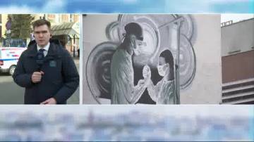 Mężczyzna w garniturze i ciemnej kurtce stoi na zewnątrz, trzymając mikrofon. Obok niego znajduje się zdjęcie przedstawiające malowidło ścienne z dwiema postaciami w maskach medycznych, dotykającymi się dłońmi.