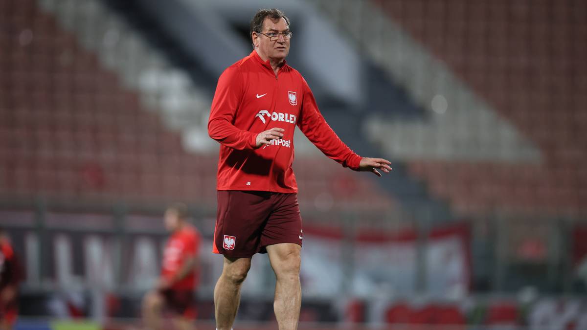 Trener w czerwonej bluzie treningowej reprezentacji Polski stoi na boisku piłkarskim.