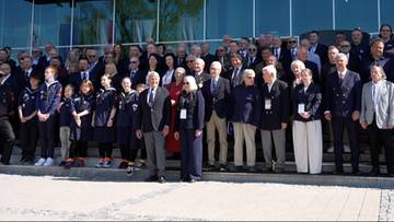 Grupa os&oacute;b w strojach sportowych i eleganckich ubraniach pozuje do zdjęcia przed nowoczesnym budynkiem.