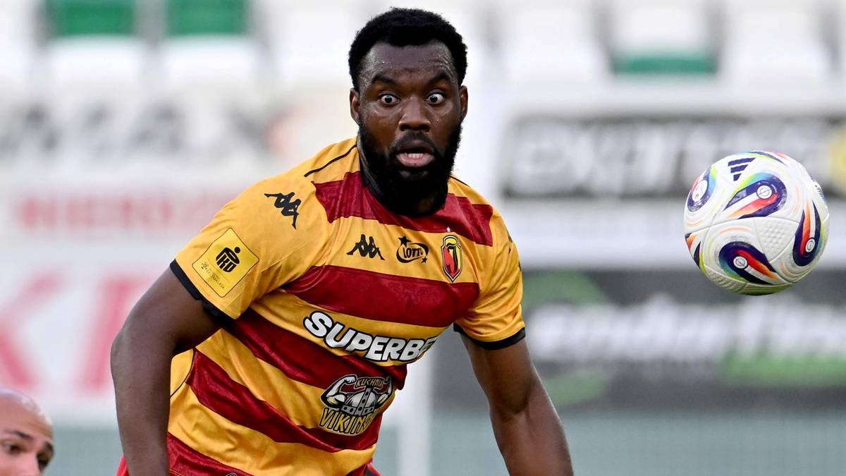 Czarnoskóry piłkarz w żółto-czerwonej koszulce grającej w piłkę nożną, z piłką obok niego, zamyślony.