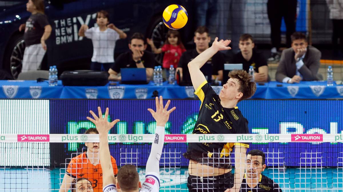 Siatkarz w ż&oacute;łto-czarnej koszulce z numerem 13 atakuje piłkę nad siatką, podczas gdy inny zawodnik w białej koszulce pr&oacute;buje go zablokować.