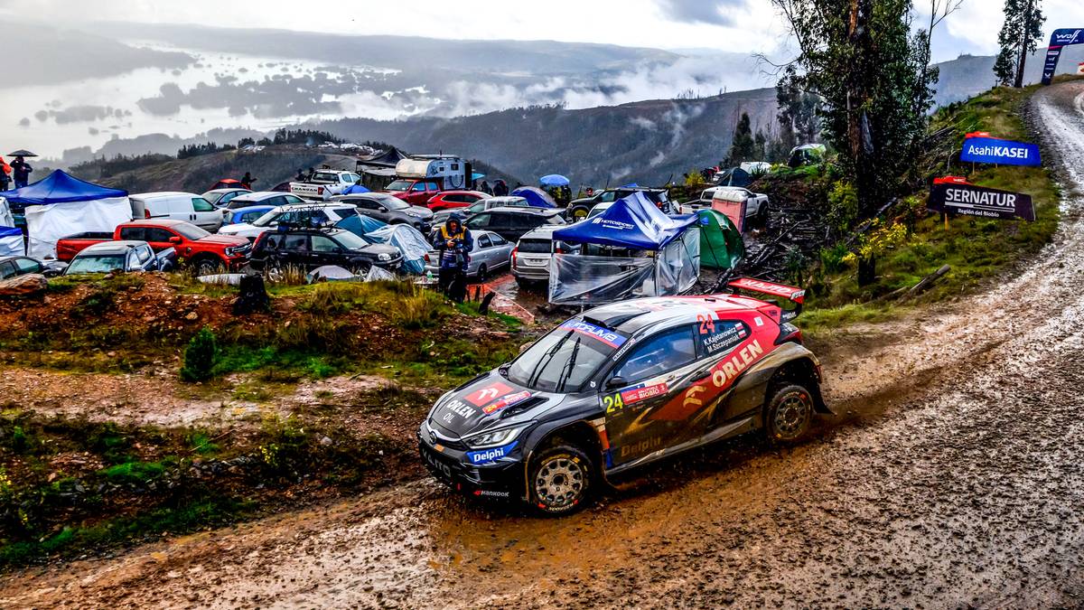 Rajdowy samochód w błocie na zakręcie toru, w tle widać widzów i pagórkowaty krajobraz.