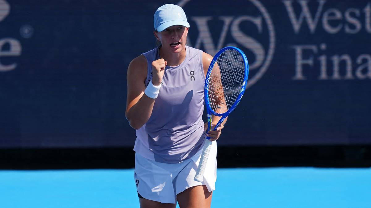 Iga Świątek, tenisistka, w szarej bluzce i białych spodenkach, w jasnoniebieskiej czapce, unosi pięść i uśmiecha się, trzymając rakietę tenisową w prawej ręce.