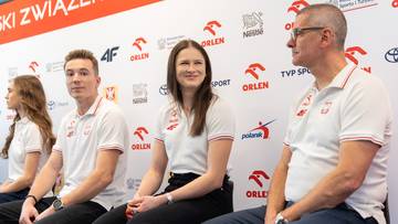 Siedzący sportowcy i trenerzy podczas konferencji prasowej, prezentujący polskie barwy narodowe.