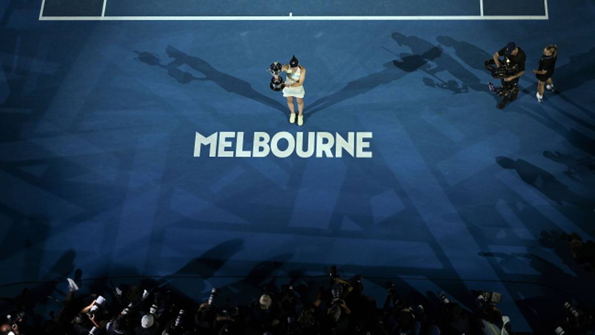 Tenisistka na niebieskim korcie z napisem "MELBOURNE" w tle, z publicznością widoczną na dole zdjęcia.