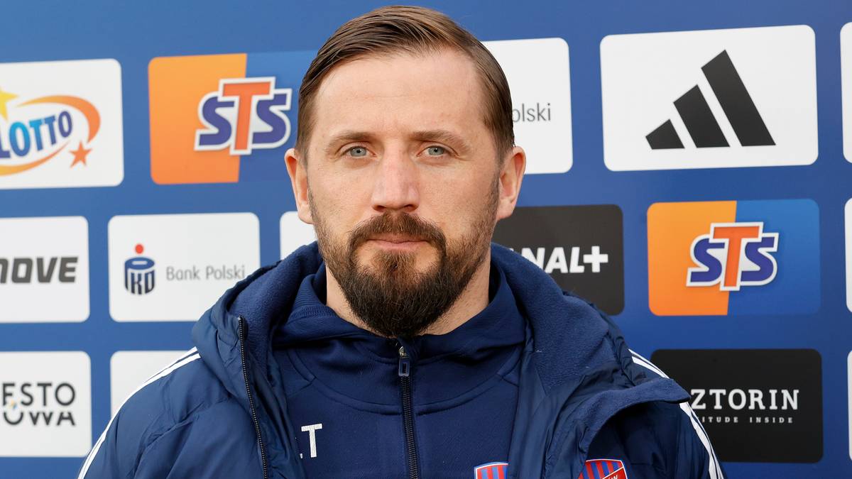 Portret mężczyzny w niebieskiej kurtce z logo Rakowa Częstochowa, stojącego przed ścianą z logotypami sponsor&oacute;w.