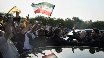 Tłum ludzi trzymający flagi Iranu, niekt&oacute;rzy unoszą ręce w geście powitania lub pozdrowienia.