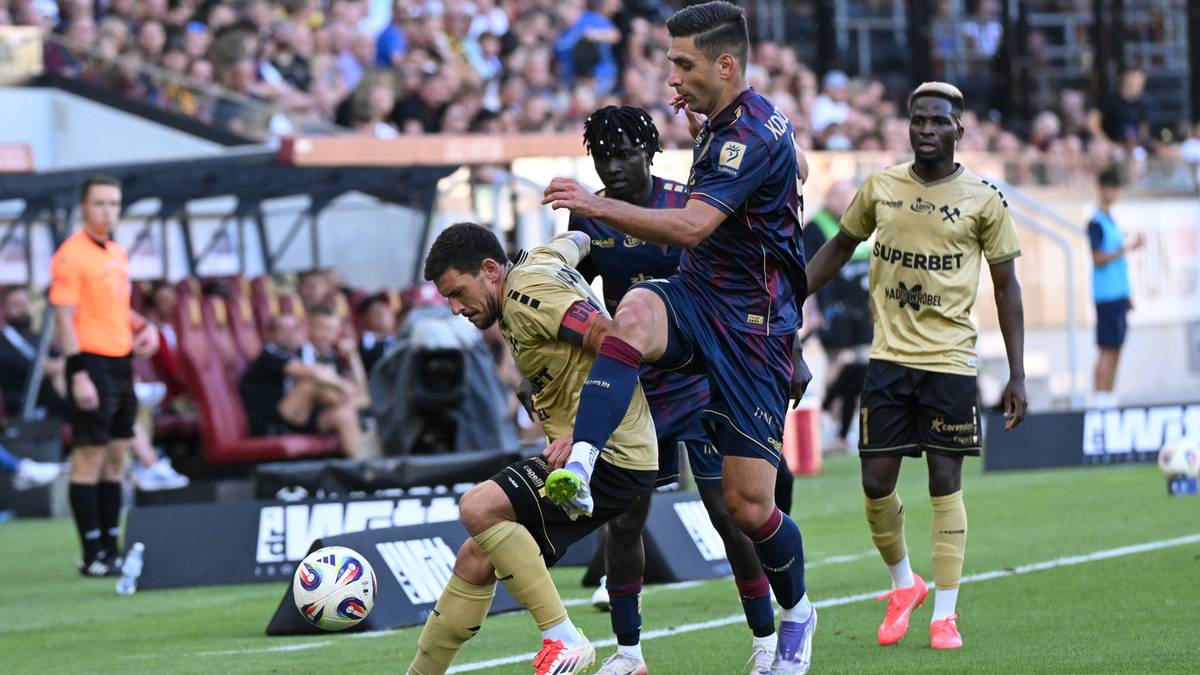 Piłkarz w żółtej koszulce podaje piłkę, podczas gdy zawodnik w niebieskiej koszulce stara się mu przeszkodzić.