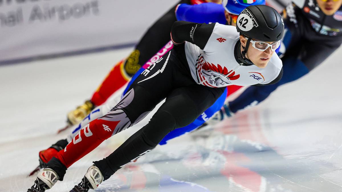 Łyżwiarz szybki w kasku i okularach ściga się na lodzie, pochylając się nisko nad taflą.