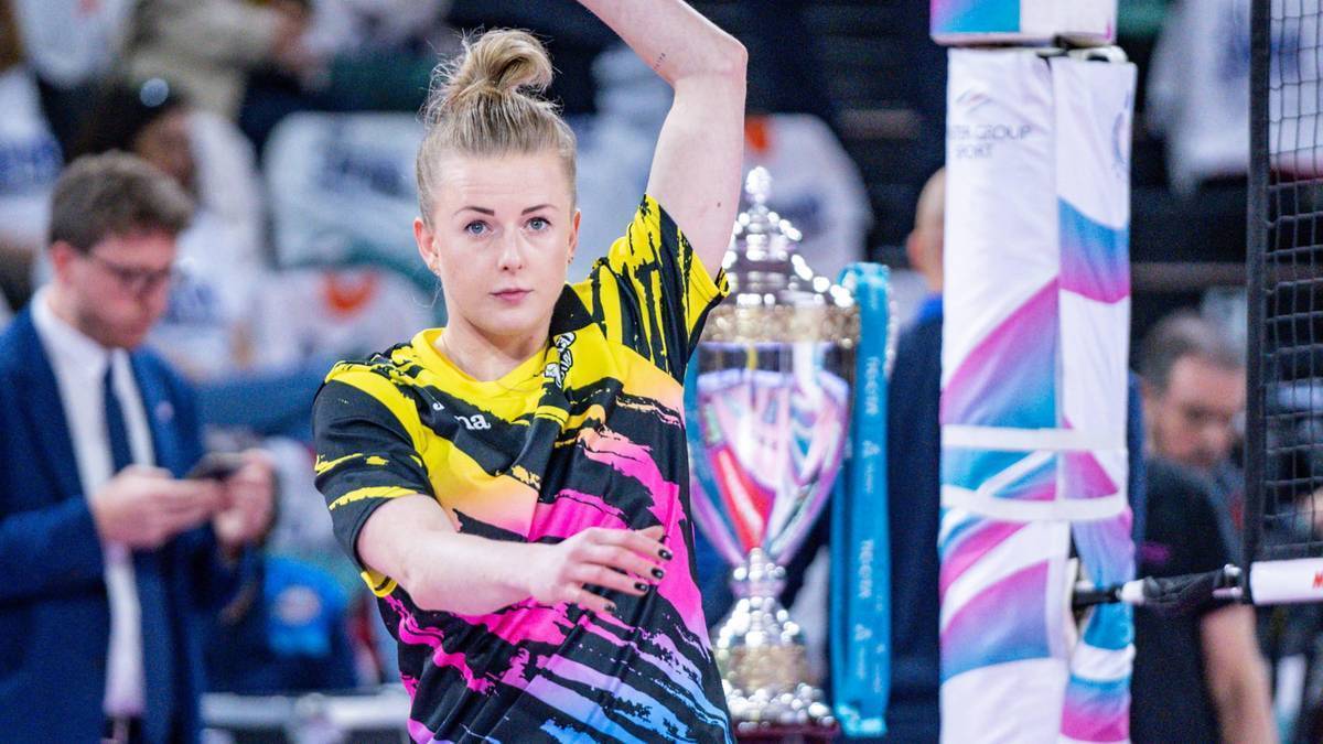 Siatkarka w kolorowej koszulce wykonuje zagrywkę, w tle widać publiczność i elementy hali sportowej.