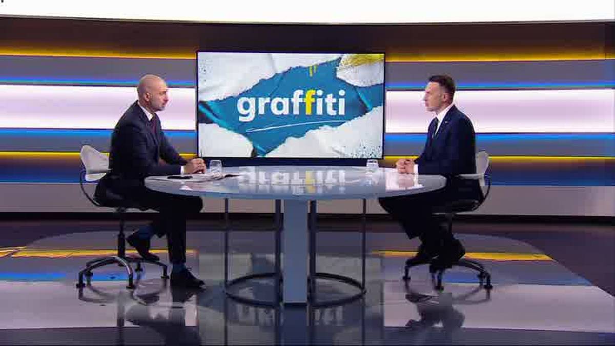 Dw&oacute;ch mężczyzn siedzących przy okrągłym stole w studiu telewizyjnym, przed nimi duży ekran z napisem "graffiti".