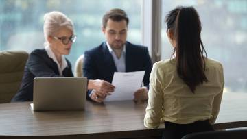 Trzy osoby siedzące przy biurku podczas rozmowy kwalifikacyjnej, dw&oacute;ch rekruter&oacute;w analizuje CV, a kandydatka odpowiada.