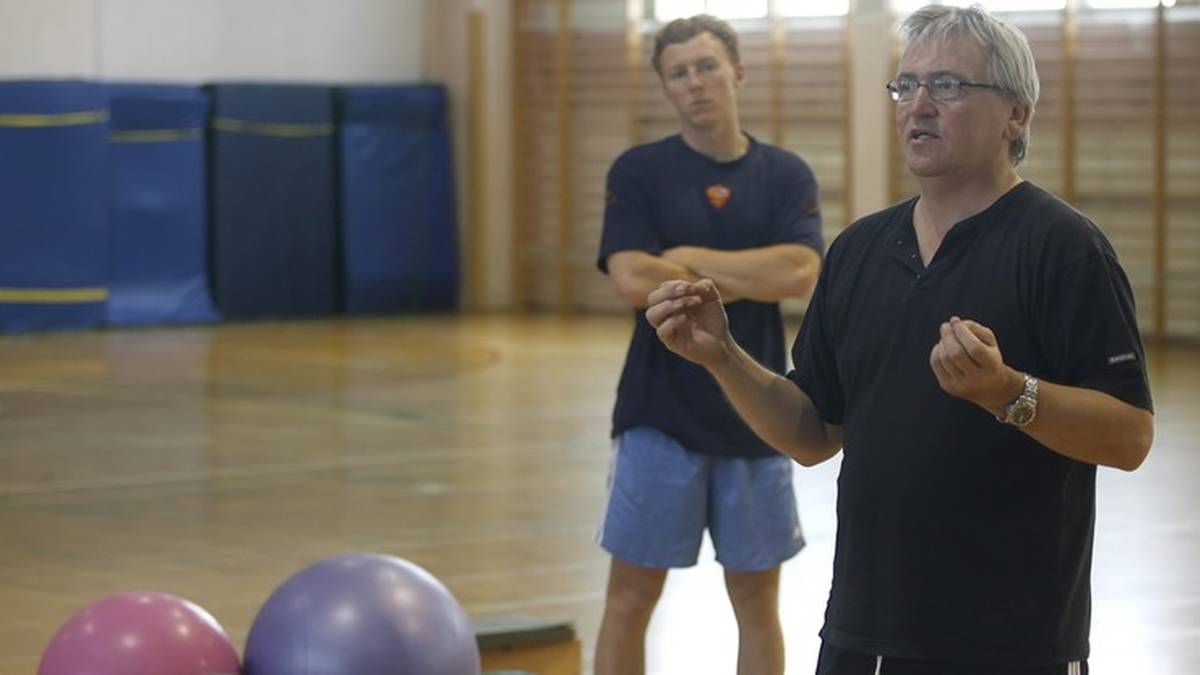 Starszy mężczyzna w okularach przemawia, gestykulując, podczas gdy młodszy mężczyzna stoi za nim z założonymi rękami w sali gimnastycznej.