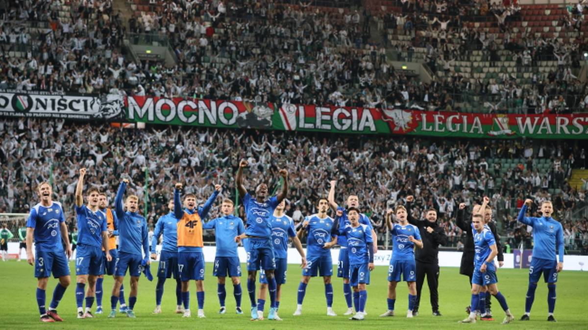 Grupa piłkarzy w niebieskich strojach świętuje na boisku piłkarskim przed trybunami wypełnionymi kibicami.