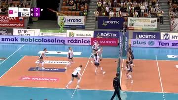 Mecz siatk&oacute;wki kobiet, zawodniczki jednej z drużyn wykonują zagrywkę, podczas gdy pozostałe przygotowują się do obrony. Na ekranie wyświetlany jest wynik meczu.