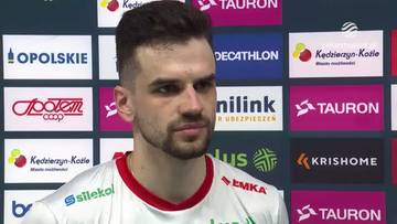 Portret Jakuba Szymańskiego, siatkarza z kr&oacute;tką brodą i ciemnymi włosami, w białej koszulce z czerwonymi elementami. Za jego plecami widać ściankę z logotypami sponsor&oacute;w, m.in. Tauron, Decathlon i Kędzierzyn-Koźle.