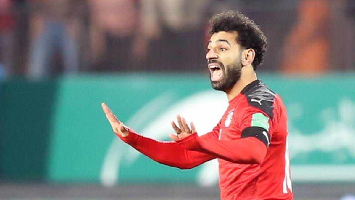 Mohamed Salah, piłkarz reprezentacji Egiptu, w czerwonej koszulce z czarnymi elementami, gestykuluje rękami na tle zielonej bandy stadionu.