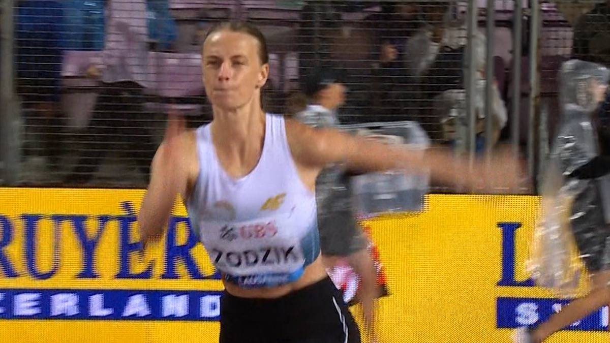 Lekkoatletka w białej koszulce sportowej z widocznym napisem "SZCZOCZ" wykonuje ruch rękami w geście triumfu, patrząc przed siebie z determinacją. Tło stanowi rozmyty tłum widzów i elementy stadionu z żółtym banerem.