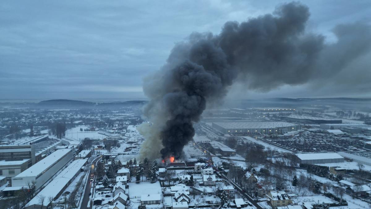 Widok z lotu ptaka na dużą halę magazynową objętą ogniem, z której wydobywa się wysoki słup czarnego dymu, unoszący się nad zabudowaniami mieszkalnymi i przemysłowymi w zimowy dzień.