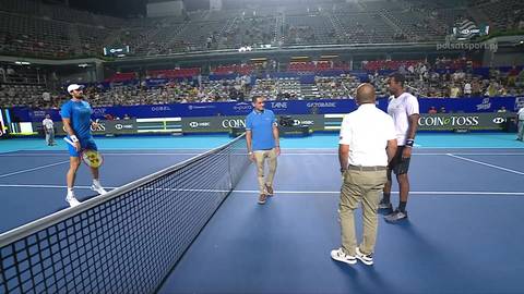 Czterech mężczyzn na korcie tenisowym, dw&oacute;ch w strojach sportowych po obu stronach siatki, dw&oacute;ch w strojach casualowych stojących obok, trybuny widowni w tle.