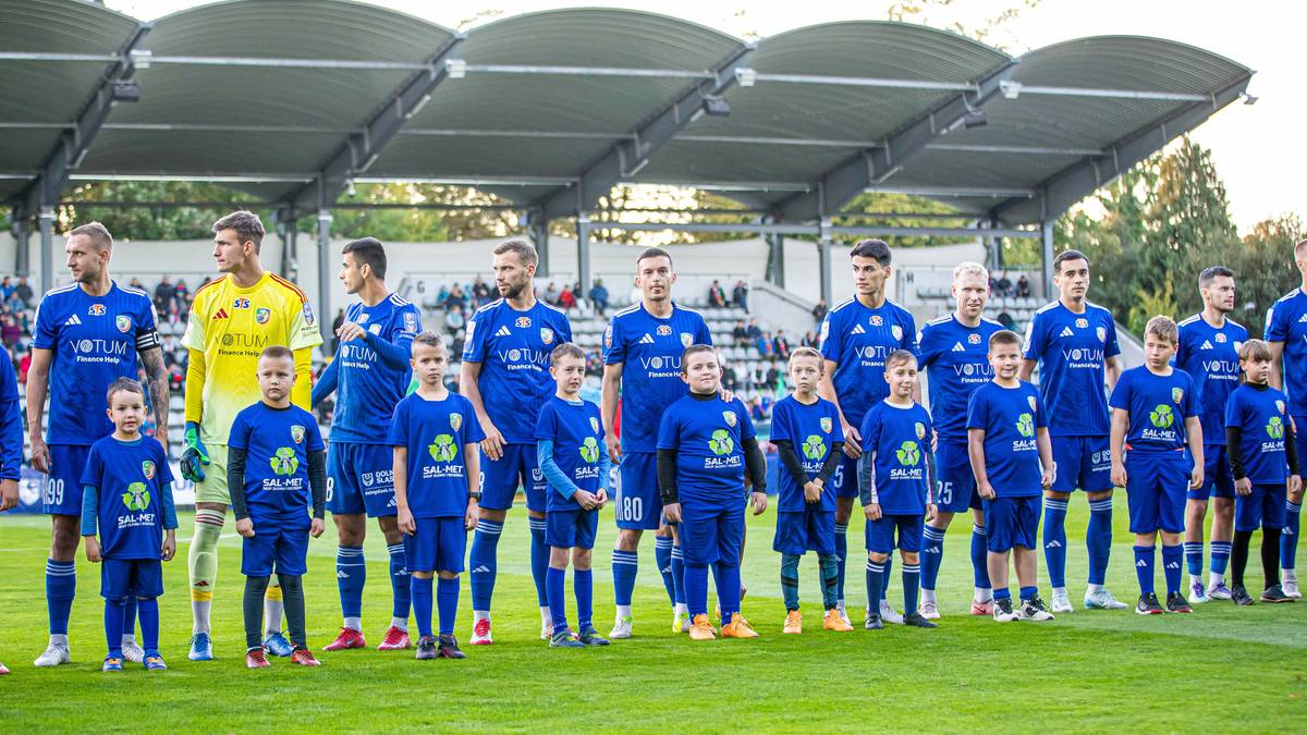 Drużyna piłkarska w niebieskich strojach, w tym młodsi zawodnicy w pierwszym rzędzie, stoi w szyku na boisku piłkarskim.