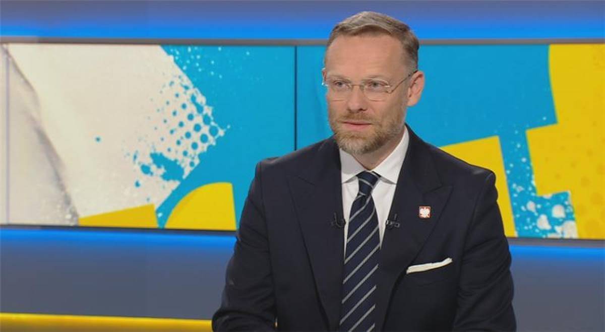 Mężczyzna w okularach i garniturze siedzi w studiu telewizyjnym.