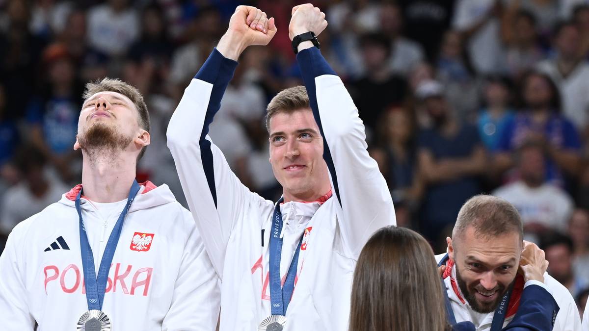 Dw&oacute;ch siatkarzy z medalami na szyjach unosi ręce w g&oacute;rę, trzeci siatkarz obok nich wita się z kimś.