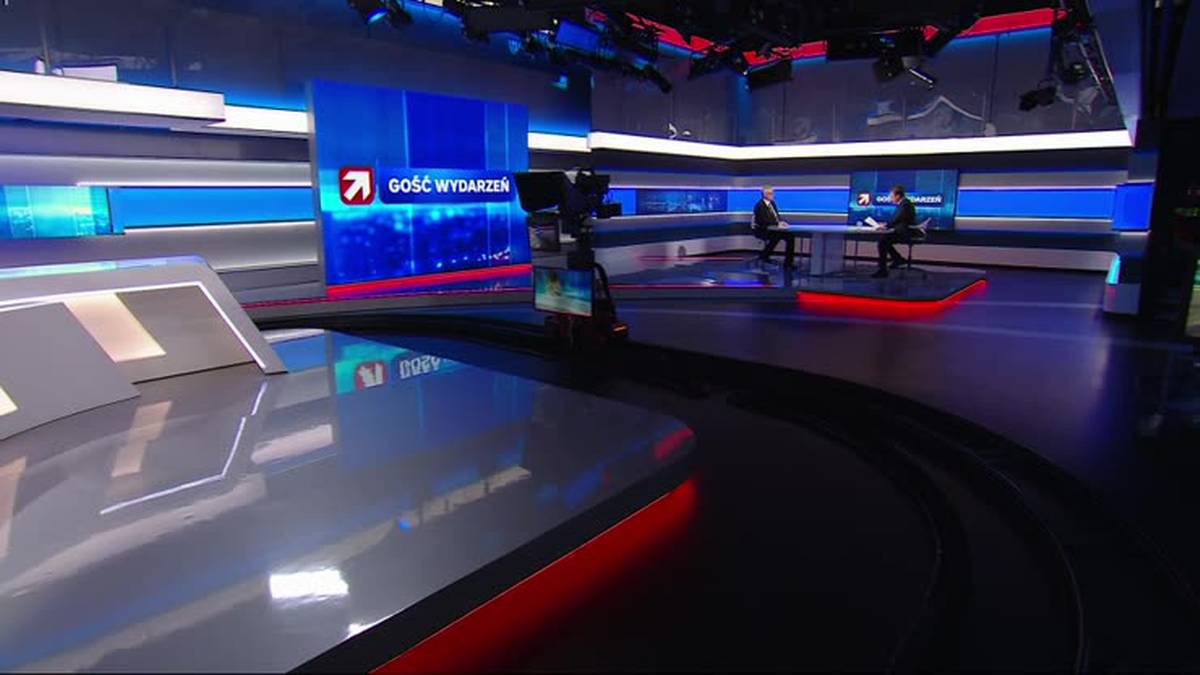 Studio telewizyjne z nowoczesnymi elementami wystroju, w tym dużymi ekranami z logo "Gość Wydarzeń". Widać r&oacute;wnież st&oacute;ł prezydialny z dwoma siedzącymi osobami.