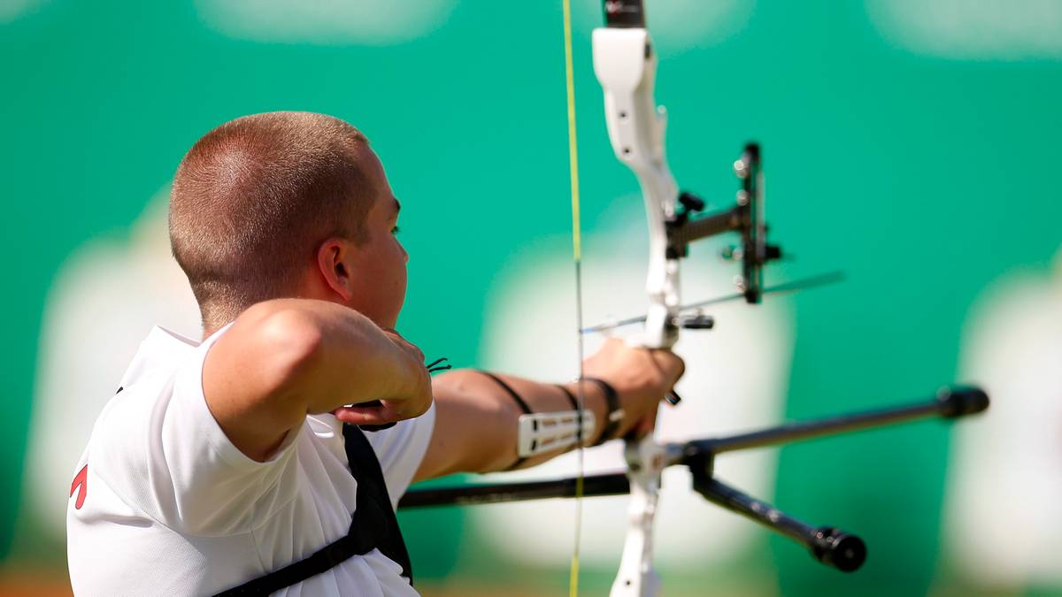 Mężczyzna w białej koszulce celuje z łuku i strzały w stronę celu.