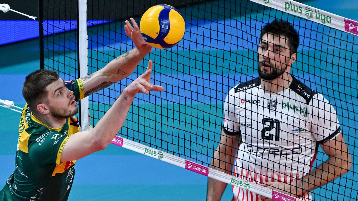 Siatkarz w zielonej koszulce wykonuje zagrywkę nad siatką, a siatkarz w białej koszulce z numerem 21 obserwuje akcję.