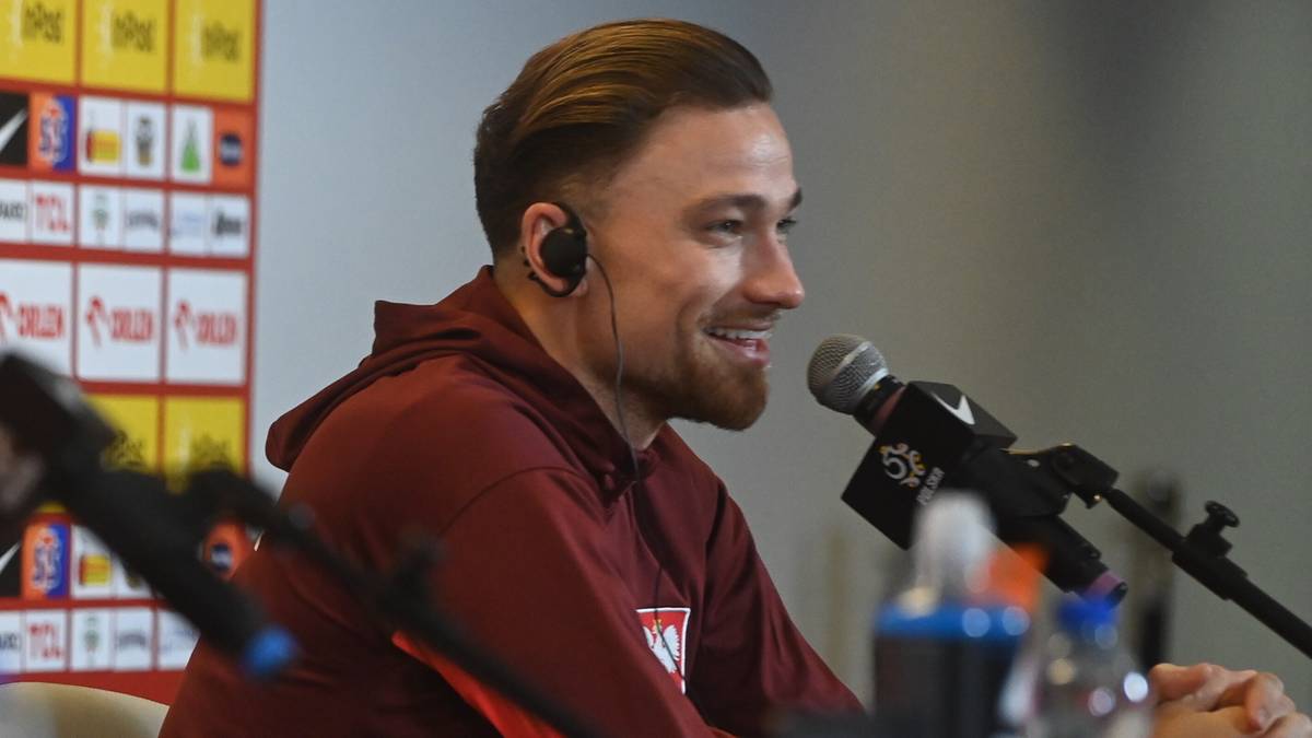 Piłkarz w bordowej bluzie z kapturem, słuchawkami na uszach, uśmiecha się, mówiąc do mikrofonu.