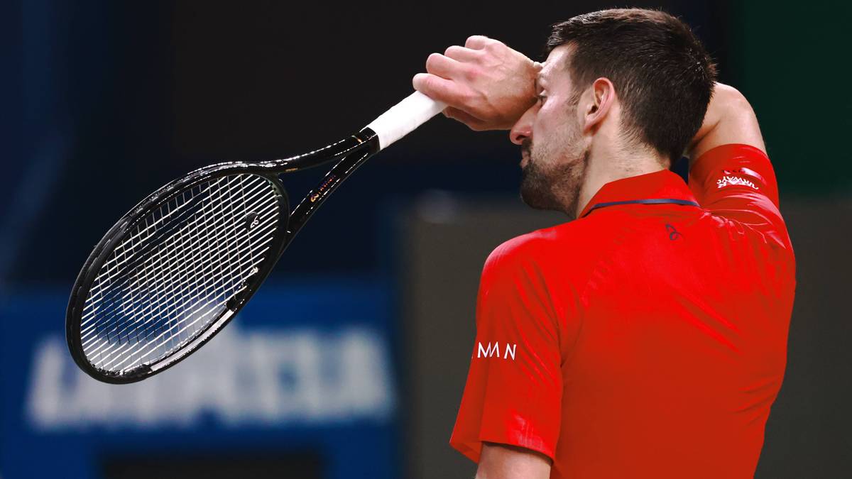 Novak Djoković w czerwonej koszulce tenisowej, trzymający rakietę tenisową nad czołem, zamyślony.