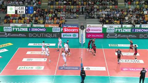 Drużyny siatkarskie w akcji na boisku podczas meczu. Widoczni są zawodnicy w białych i czerwonych strojach, a także sędzia przy siatce. Na trybunach zgromadzeni są kibice. Na ekranie widoczny jest wynik "CZE 0 1 RZE".