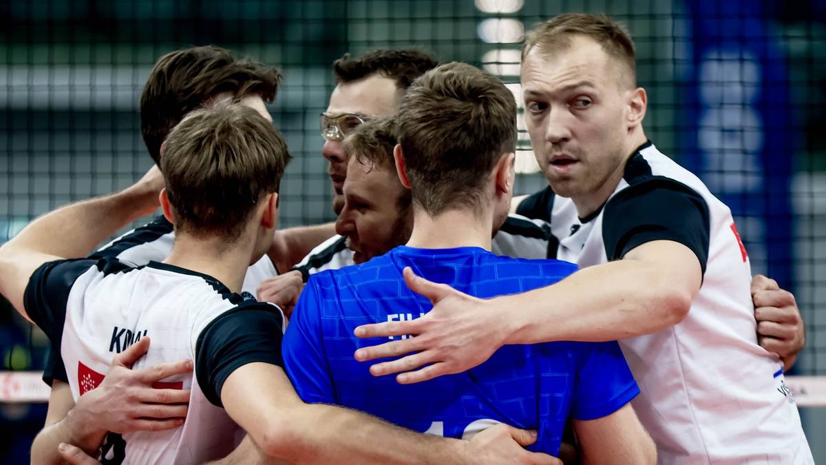 Grupa siatkarzy w białych i niebieskich strojach przytulających się w geście wsparcia na boisku siatkarskim.