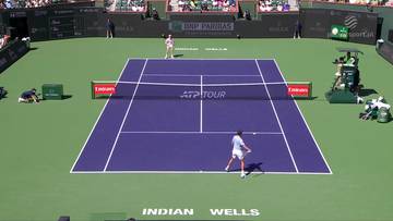 Dwóch tenisistów gra mecz na korcie tenisowym podczas turnieju ATP Tour. Na korcie widać dwóch zawodników, jeden z nich właśnie uderza piłkę rakietą. W tle znajdują się trybuny z widzami oraz reklamy sponsorów.