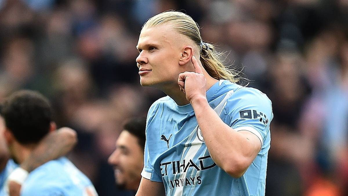 Piłkarz Erling Haaland w jasnoniebieskiej koszulce Manchesteru City z numerem 9 na plecach, odwr&oacute;cony w prawo, z ręką na szyi, na tle innych piłkarzy i publiczności.