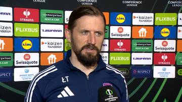 Łukasz Tomczyk, trener, stoi na tle ściany ze sponsorami.