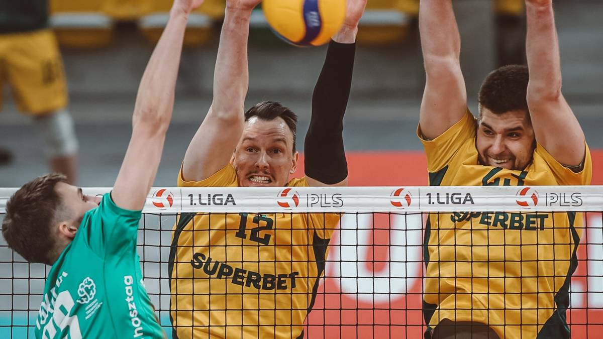 Trzech siatkarzy w akcji podczas meczu, dw&oacute;ch w ż&oacute;łtych koszulkach z numerami 12 i 10 pr&oacute;bują zablokować atak przeciwnika, kt&oacute;ry w zielonej koszulce wyskakuje do zagrania piłki nad siatką.