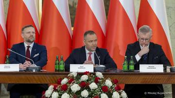 Trzech mężczyzn w garniturach siedzi przy stole podczas oficjalnego wydarzenia, na tle polskich flag.