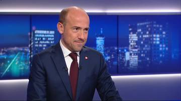 Borys Budka, ubrany w granatowy garnitur i ciemnoczerwony krawat, spogląda w prawo z lekkim uśmiechem. W tle znajduje się ekran z animowanym widokiem nocnego miasta z wieżowcami i światłami.